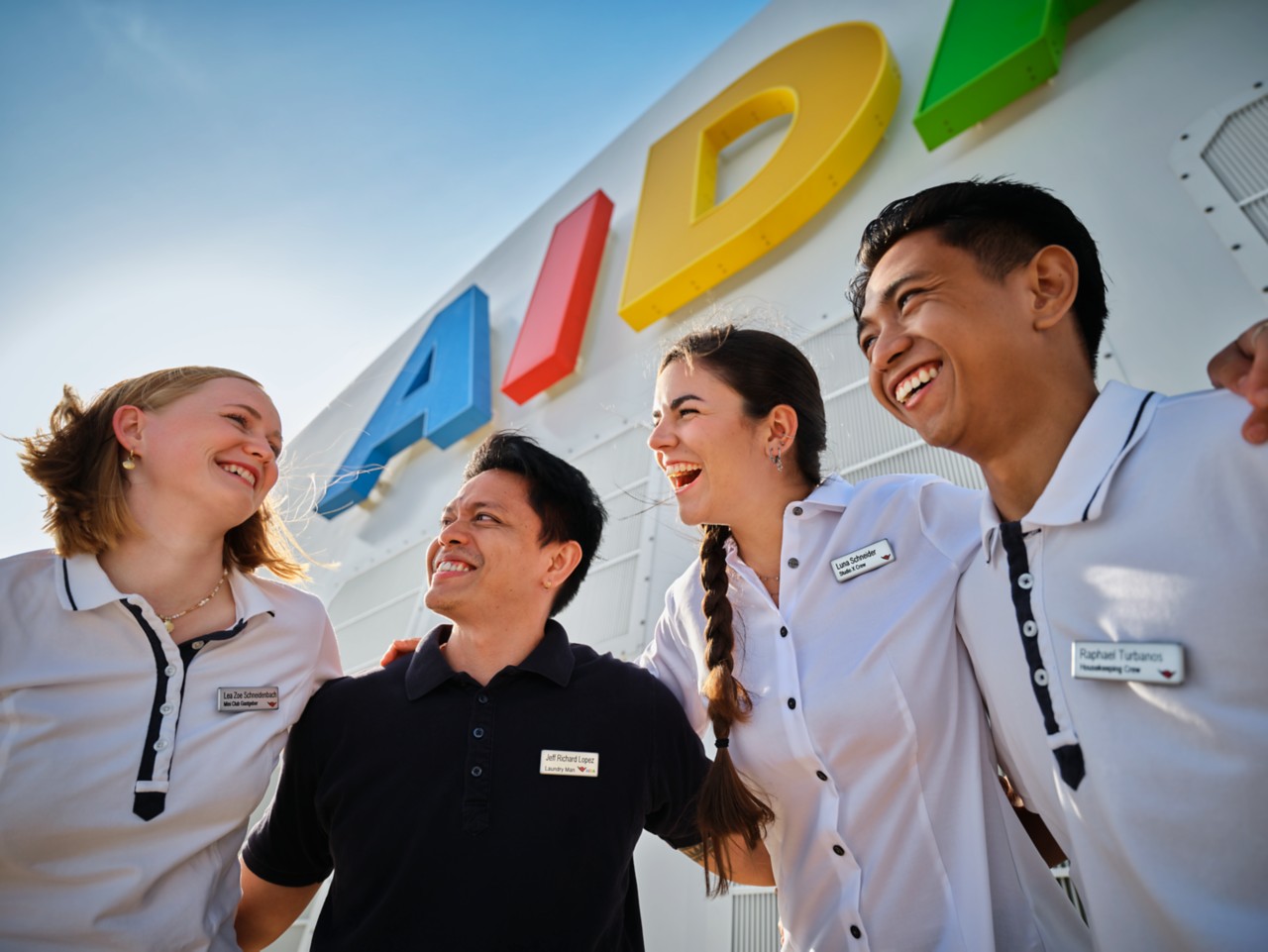 Vier lächelnde Crewmitglieder von AIDA Cruises stehen Arm in Arm vor dem AIDA Schriftzug an Bord