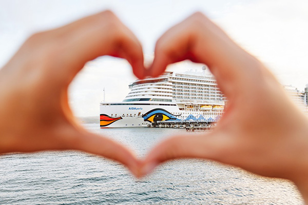 Zwei Hände formen ein Herz und rahmen ein AIDA-Kreuzfahrtschiff mit Kussmund und Auge auf dem Wasser ein.